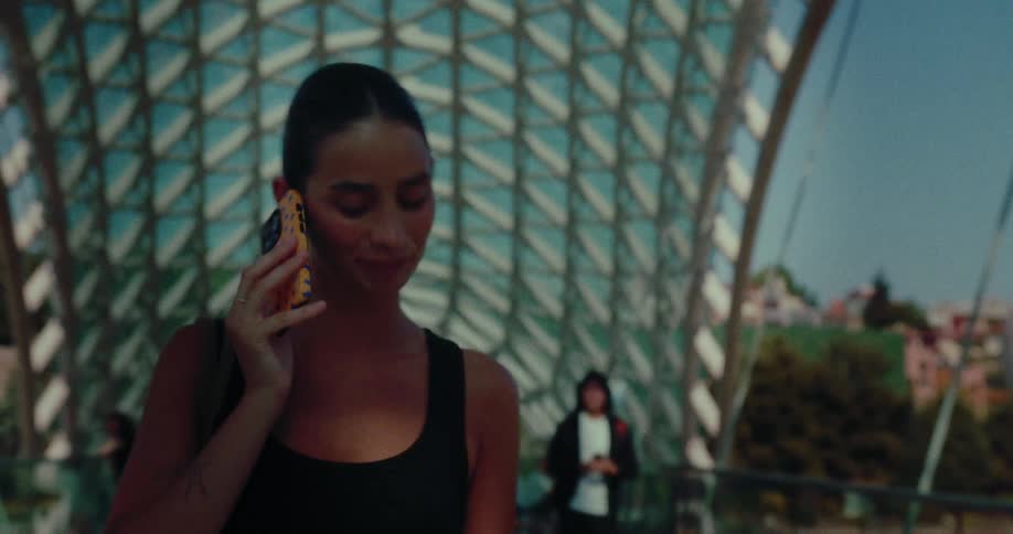 A young woman with her hair in a bun talks on her smartphone while walking across a modern glass and steel bridge on a sunny day