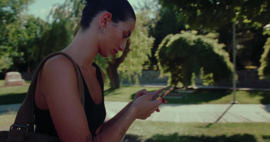 A young woman with her hair up walks through a sunny park while looking down and typing on her smartphone