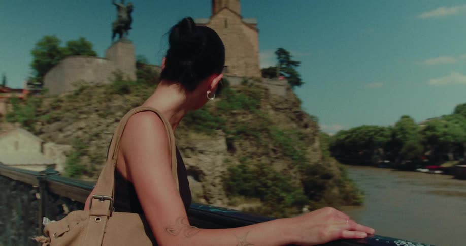 A stylish young woman gazes at the scenic view of a historic city and river from an ornate bridge on a sunny day