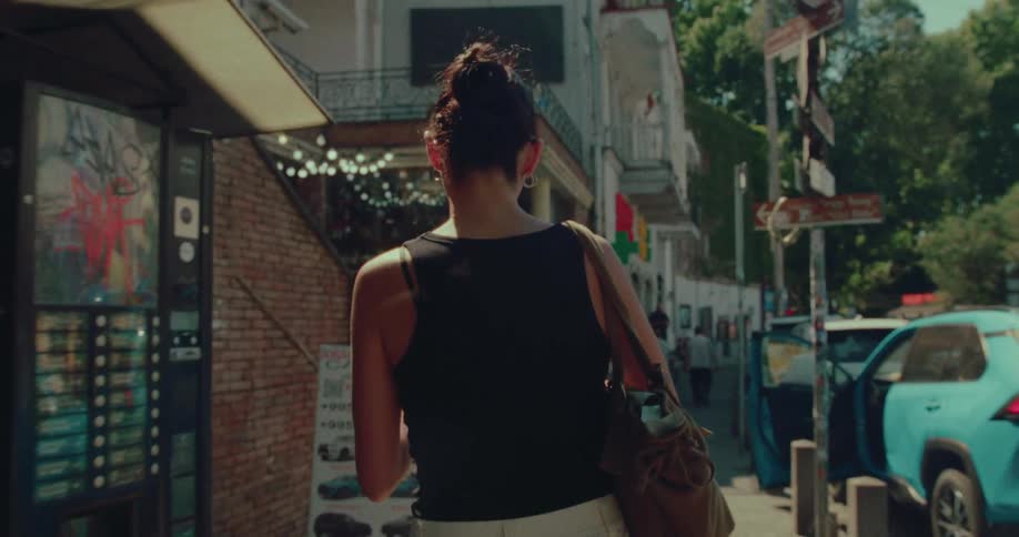 An over-the-shoulder slow-motion shot follows a woman walking down a sunny city sidewalk past cars and storefronts