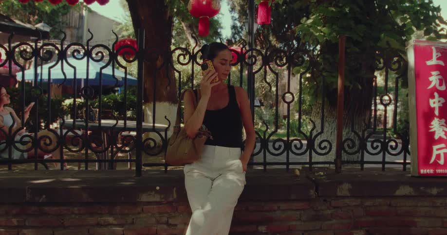 A stylish woman talks on her cell phone while leaning against a brick wall on a sunny day in an urban Asian setting
