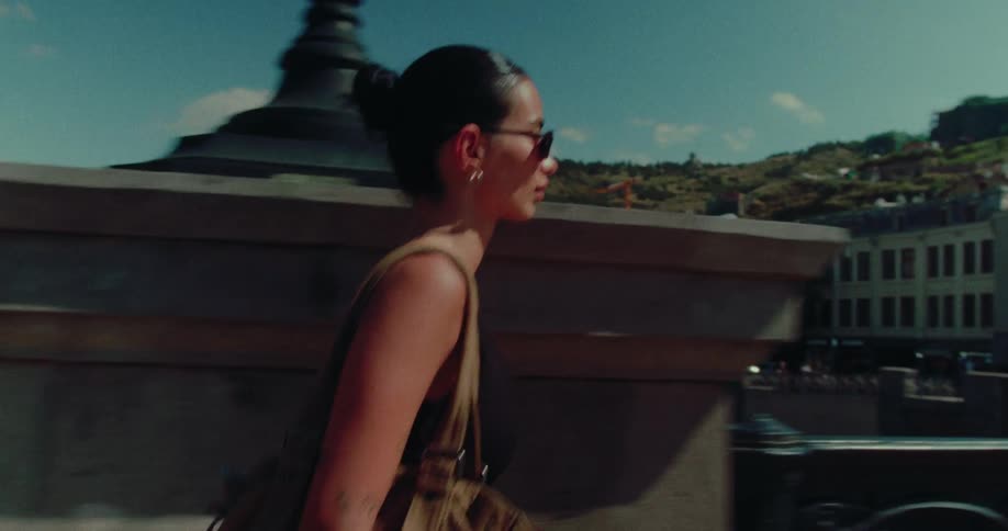 A young brunette woman in sunglasses enjoys a leisurely walk across a beautiful city bridge on a sunny summer day