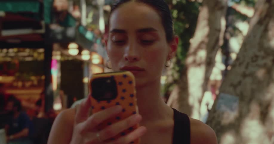 Focused young woman uses her smartphone while walking down a sunlit, tree-lined street with shops in the background
