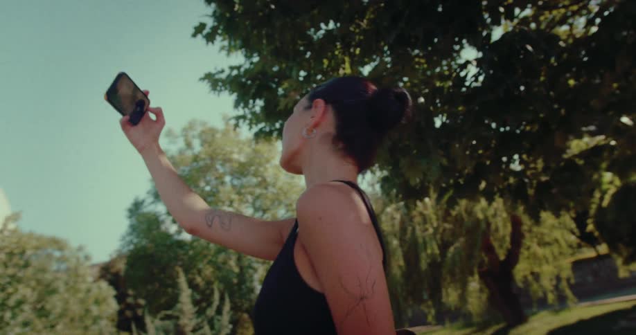A young woman in a black tank top takes a selfie while strolling through a sunny, green park