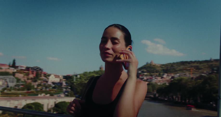 A beautiful young woman talks on her cell phone while standing on a city bridge overlooking a river on a sunny day