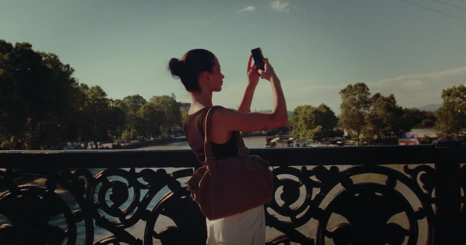A stylish female tourist captures memories on her smartphone while standing on an ornate bridge overlooking a river at sunset