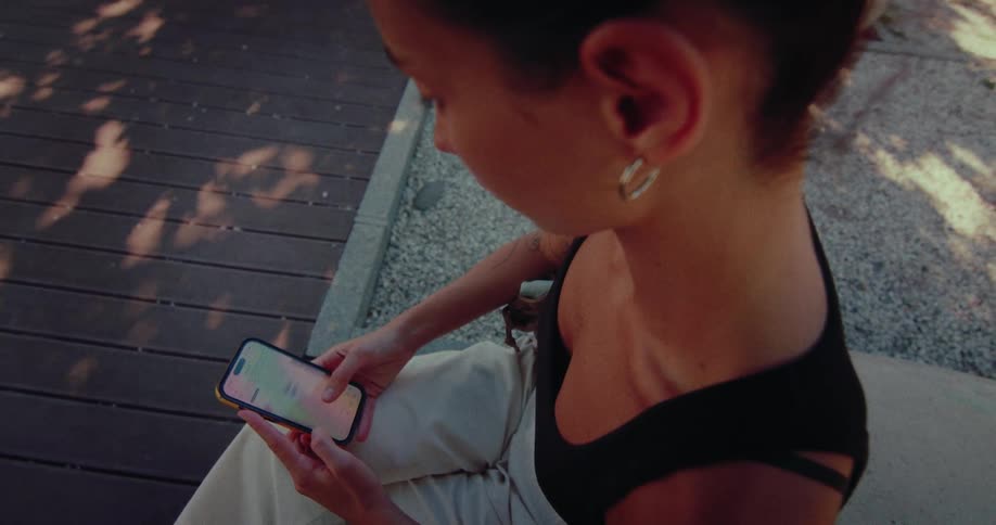 Over-the-shoulder view of a young woman sitting on a wooden bench and scrolling through her smartphone in the warm evening light