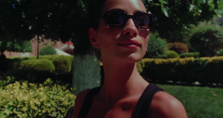 A beautiful young woman in sunglasses smiles flirtatiously at the camera in a sunny park in slow motion