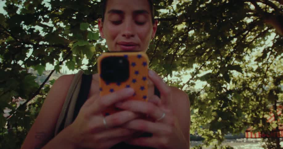 A smiling young woman reacts with surprise and happiness while using her smartphone outdoors under a tree