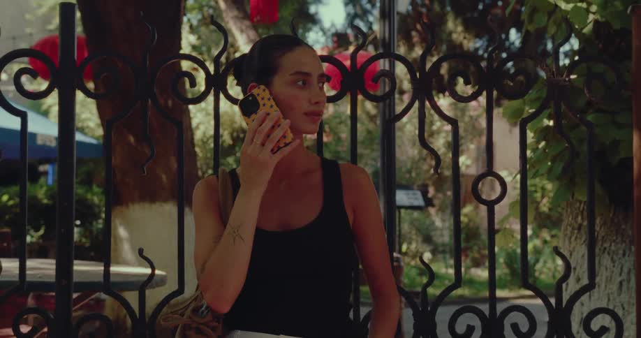 A young woman with a tattoo stands by a decorative iron gate on a sunny day while engaged in a phone conversation