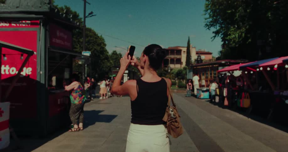 A woman captures memories on her smartphone while walking down a bustling market street in Georgia on a sunny day