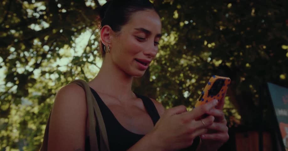 A stylish young woman smiles while using her smartphone outdoors under the shade of a leafy tree on a sunny day