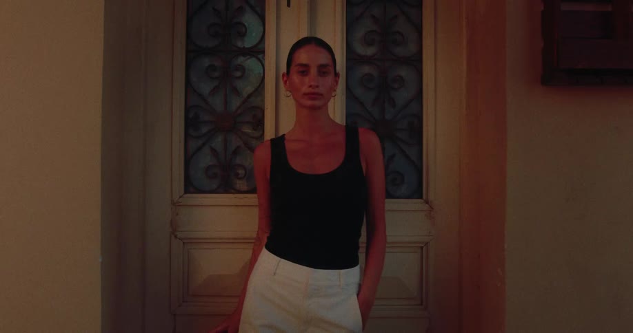 A fashionable woman takes off her sunglasses and poses with confidence in a warmly lit doorway at night