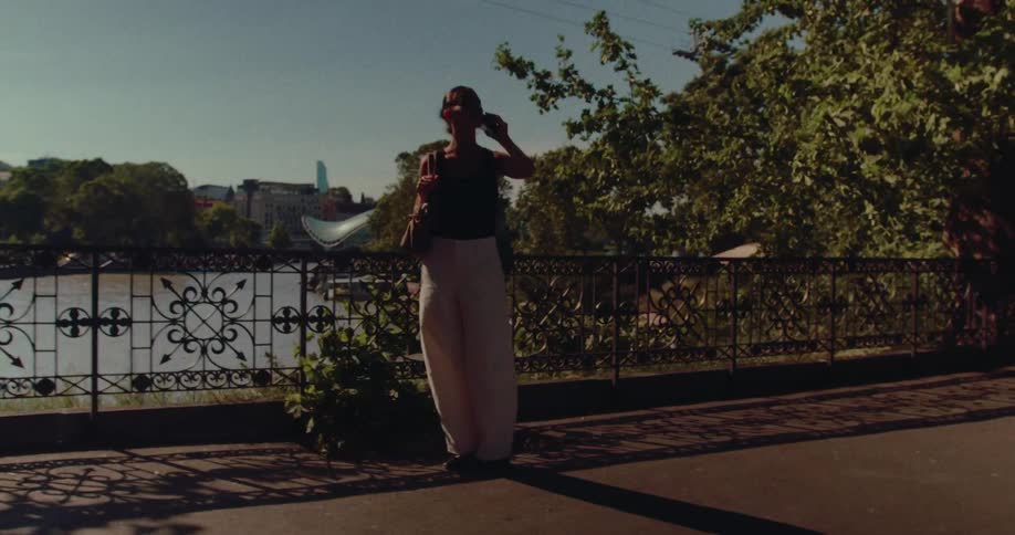 A stylish woman walks along a sunny, tree-lined riverside promenade while talking on her phone in the city