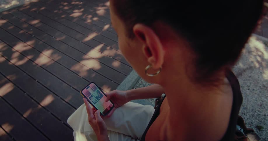 High-angle, over-the-shoulder view of a woman scrolling through a social media feed on her smartphone while sitting outdoors