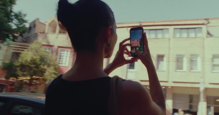 A young woman stands on a sunny city sidewalk, using her smartphone to photograph the local architecture while exploring