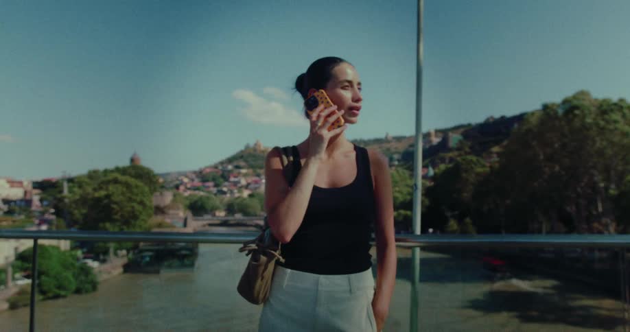 A stylish young woman talks on her phone while standing on a modern bridge overlooking a river and a scenic old town on a sunny day
