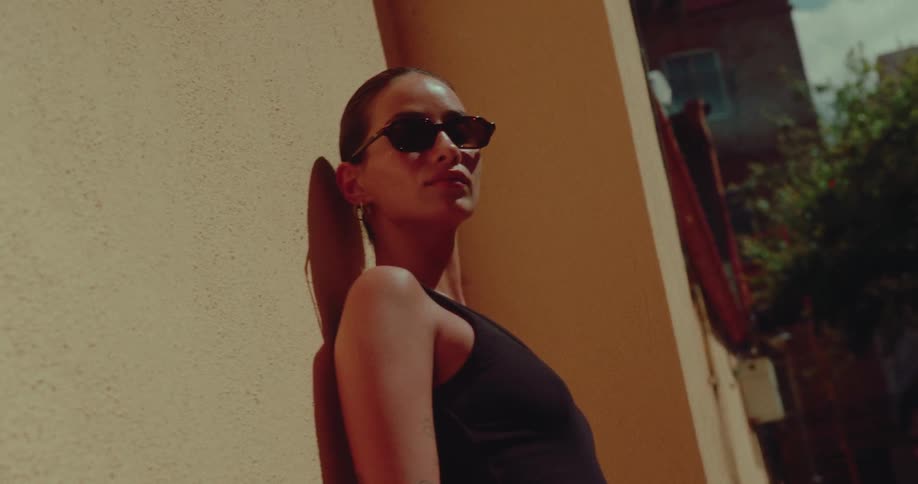 A stylish woman in sunglasses leans against a sun-drenched yellow wall, basking in the warm afternoon light