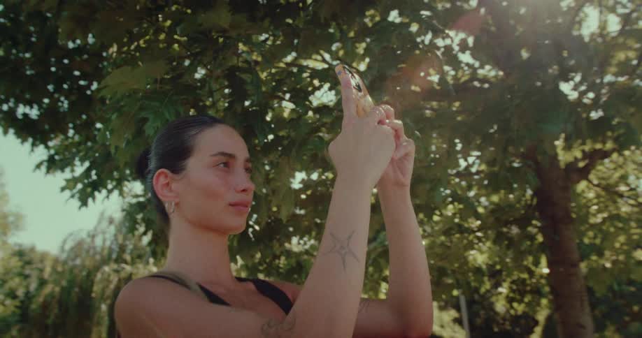 A young woman with tattoos stands under a large tree in a sunny park, taking a photo with her smartphone
