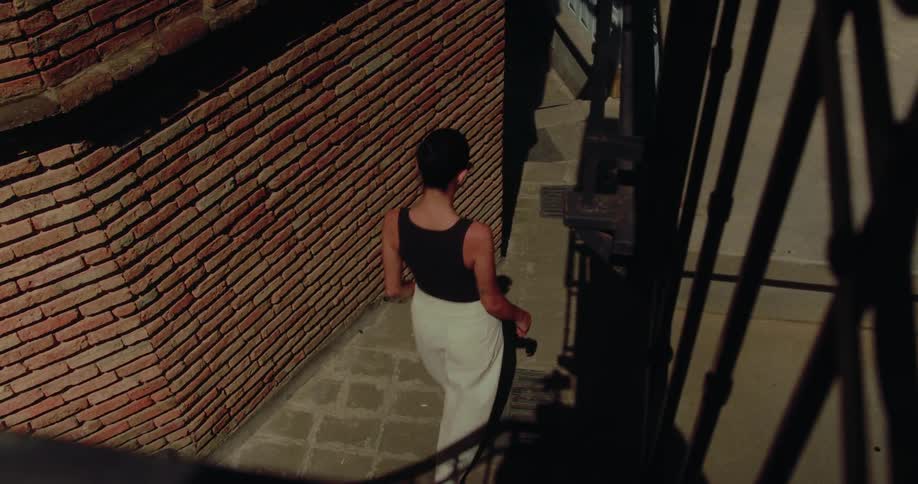 High-angle slow motion shot of a woman walking down sunlit stairs in a narrow brick alleyway