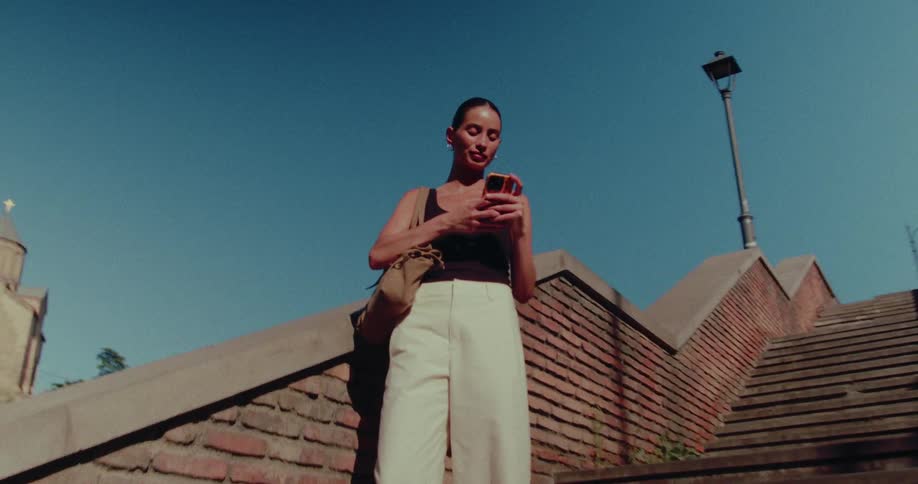 A stylish woman stands on a brick staircase on a sunny day, engrossed in her smartphone