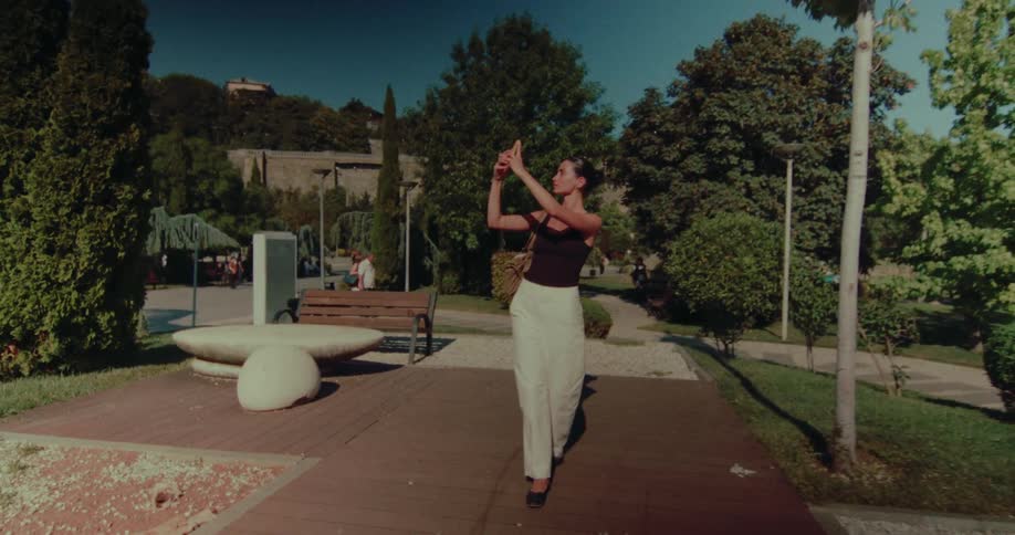 A young woman walks backwards through a sunny urban park, capturing the moment on her smartphone