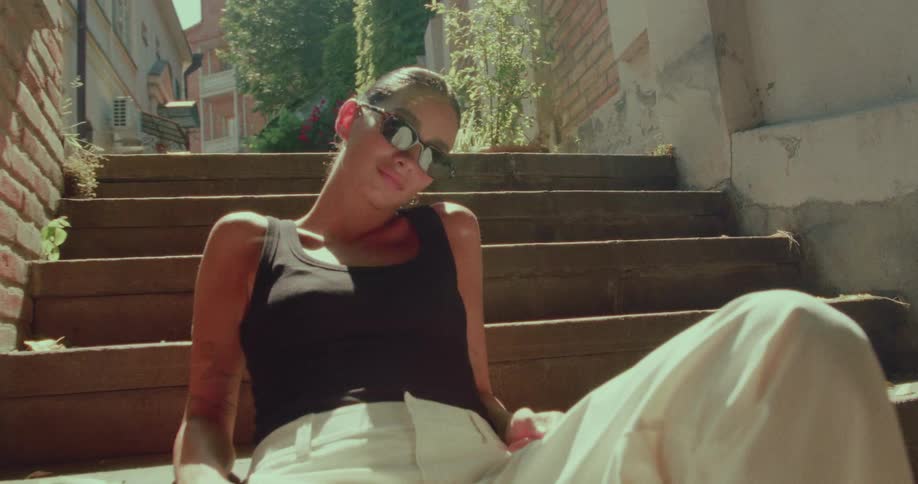 A stylish young woman wearing sunglasses poses casually while sitting on old wooden stairs in a sunny city alley