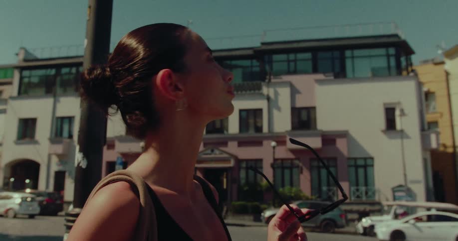 A stylish woman takes off her sunglasses and enjoys the summer sun while standing on a bustling city street in slow motion