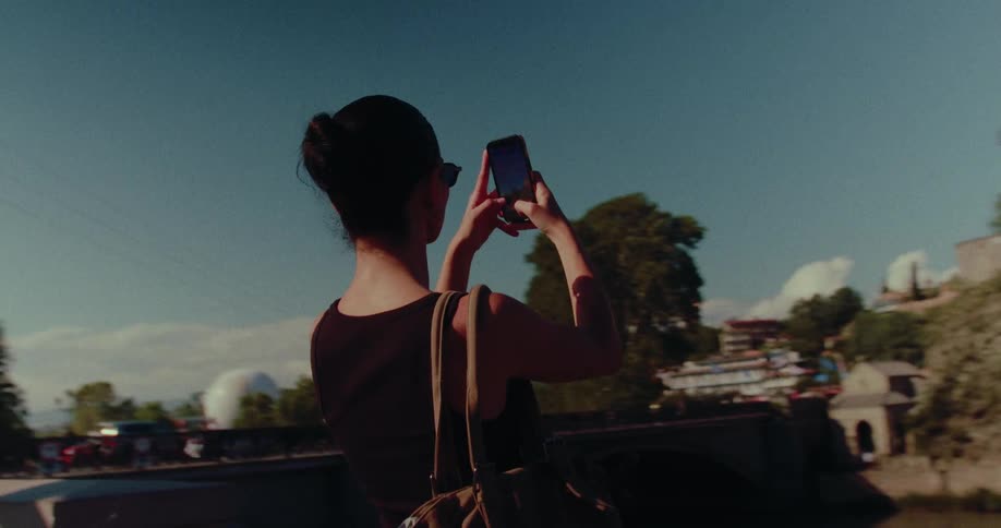 A female tourist stands on a bridge, capturing video of the historic cityscape and Metekhi Church in Tbilisi, Georgia with her smartphone