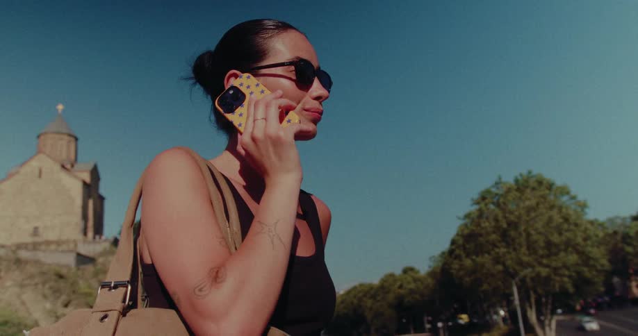 Stylish woman in sunglasses answers a call while walking on a bridge in front of an ancient European church on a sunny day