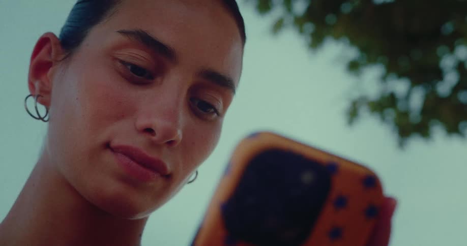 A low-angle close-up shot of a young woman smiling as she looks down at her smartphone while relaxing outdoors