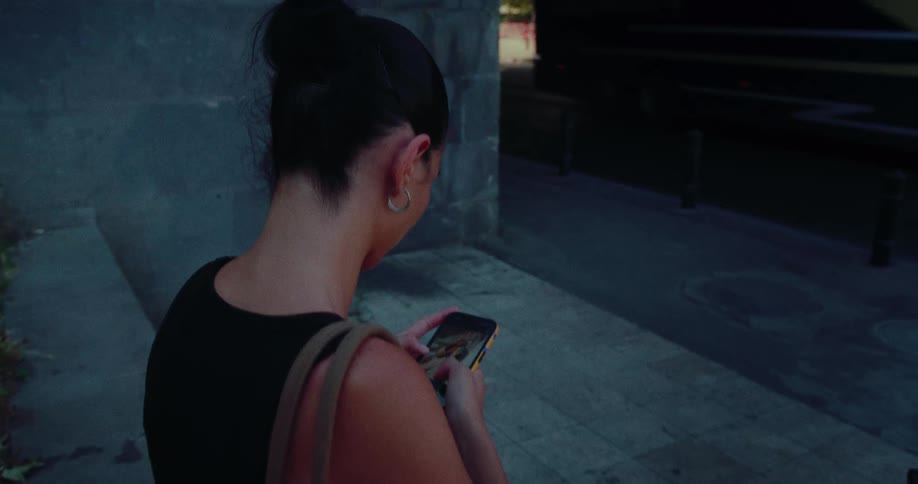 A woman scrolls on her smartphone while standing on a city sidewalk at night with traffic passing by in the background