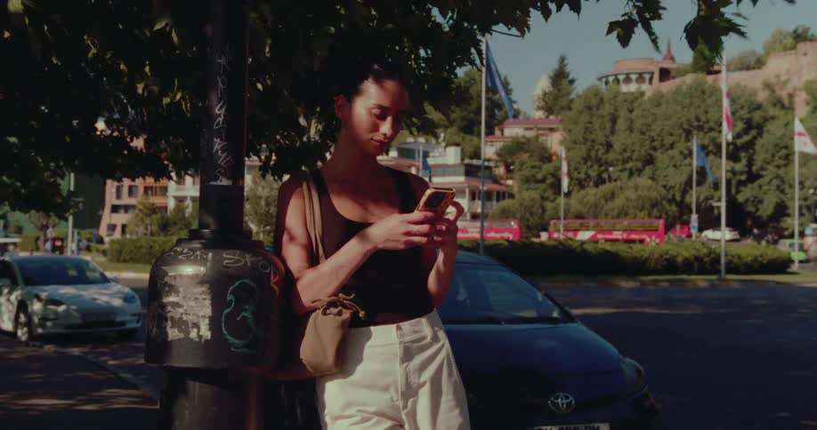 A stylish woman stands in the shade of a tree on a sunny city street, leaning against a lamp post while using her smartphone
