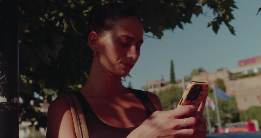 A beautiful young woman checks her phone while standing in the shade of a tree in a European city on a sunny day