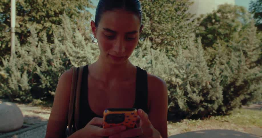 The camera slowly pushes in on a stylish young woman sitting on a contemporary stone bench in a sunlit park, absorbed in her smartphone