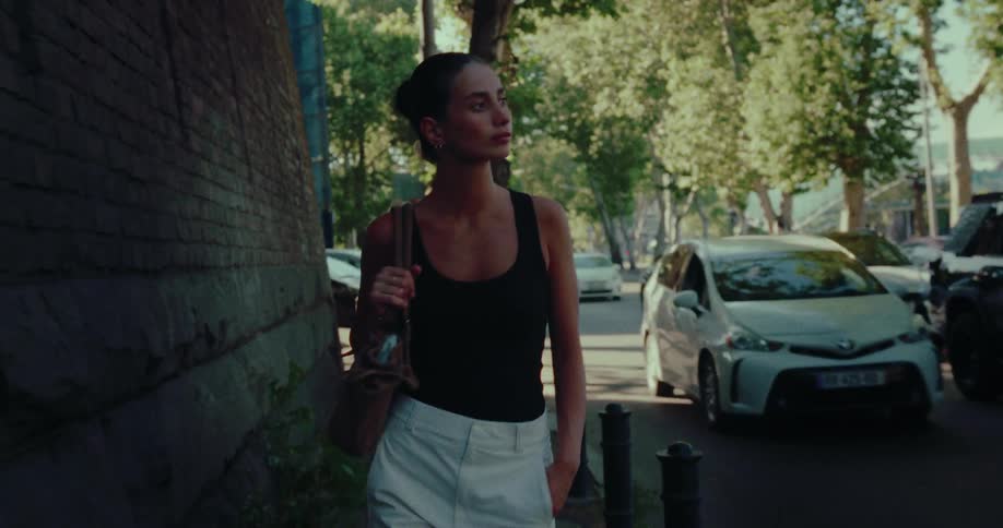 A stylish woman walks in slow motion along a sun-dappled city sidewalk next to traffic