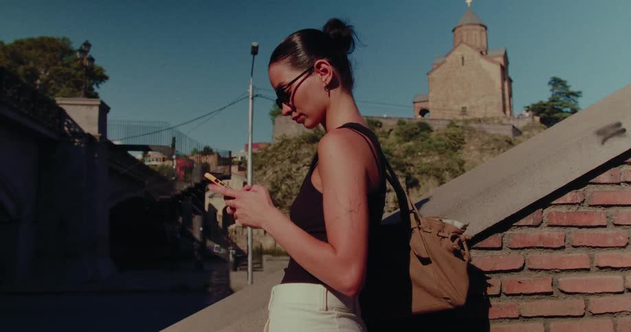 A stylish woman in sunglasses scrolls on her smartphone on a sunny day with a historic church in the background