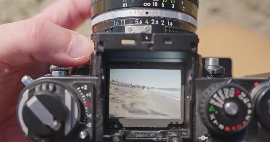 Close-up of a hand focusing a vintage SLR camera, with a sunny beach and ocean waves visible in the viewfinder