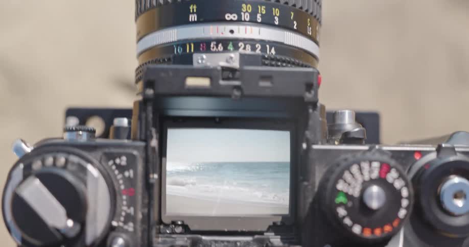 A classic film camera's viewfinder frames a beautiful scene of ocean waves rolling onto a sandy shore
