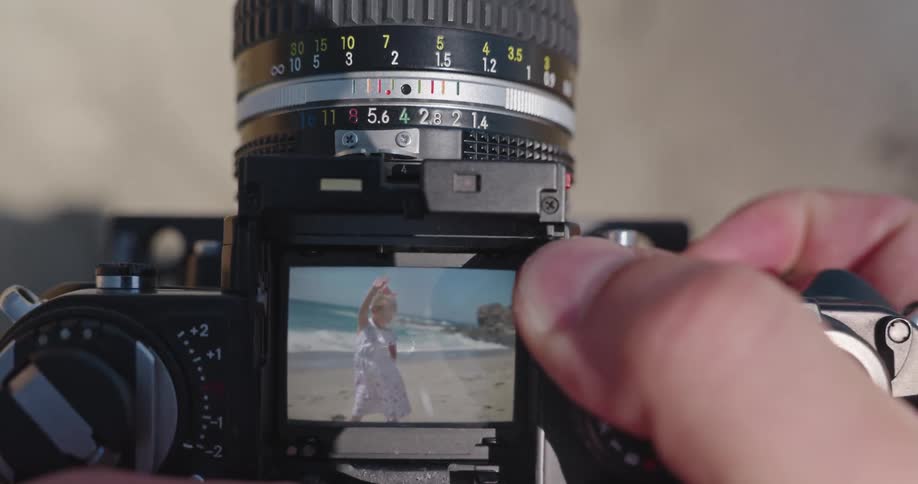 Close up of a photographer adjusting the focus ring of a vintage camera, capturing a little girl waving on the beach as seen through the viewfinder