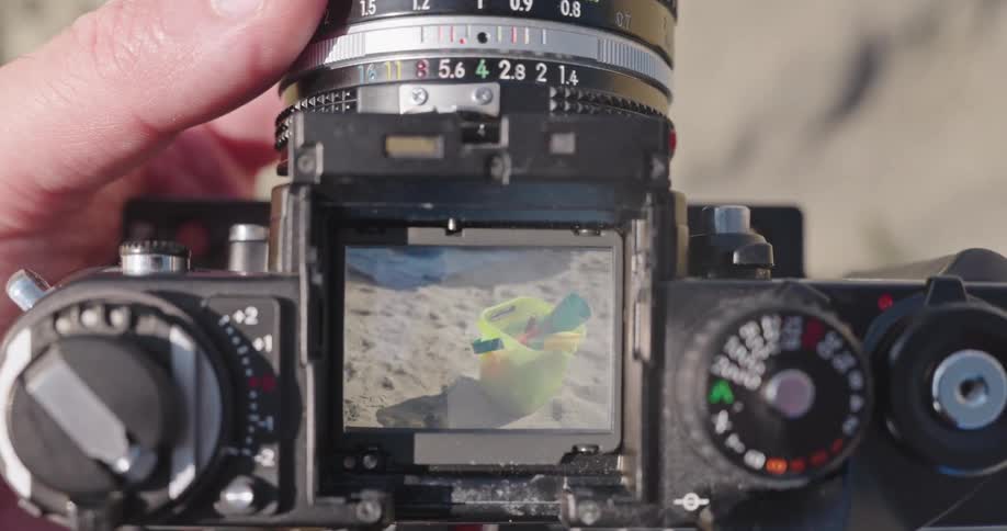 Looking through the viewfinder of a vintage SLR camera while focusing on a colorful bucket and spade on a sandy beach