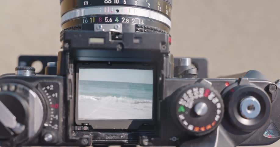 Slow-motion ocean waves wash onto the shore, as seen through the waist-level viewfinder of a classic film camera