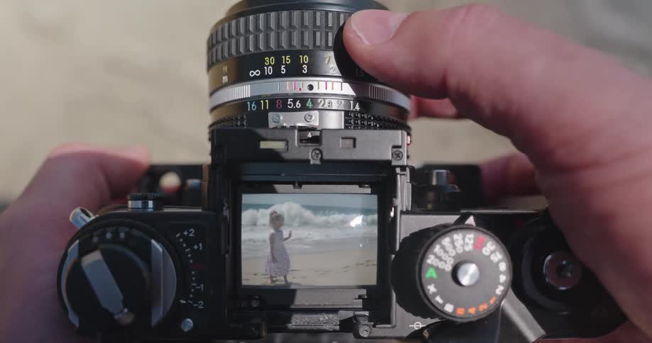 A first-person point of view of a photographer focusing a vintage film camera on a young girl playing on a sunny beach by the ocean