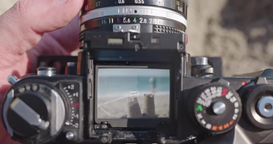 Close-up of a photographer focusing a vintage camera on a sandcastle at the beach, seen through the waist-level viewfinder
