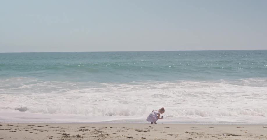 A carefree little girl in a floral dress runs toward the ocean waves on a sandy beach in slow motion