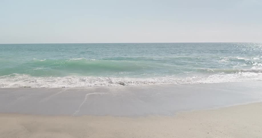 Slow motion shot of gentle turquoise ocean waves washing over a sandy beach on a bright, peaceful day