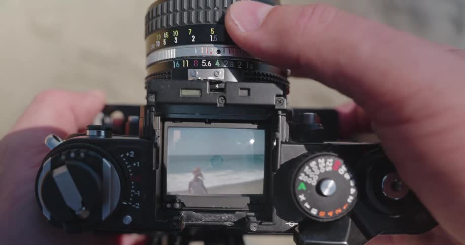 A photographer adjusts the focus on a vintage film camera, framing a shot of a young child playing on the sandy beach by the ocean