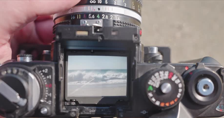 A close-up, top-down view shows a hand focusing a vintage camera on ocean waves crashing onto the shore in slow motion