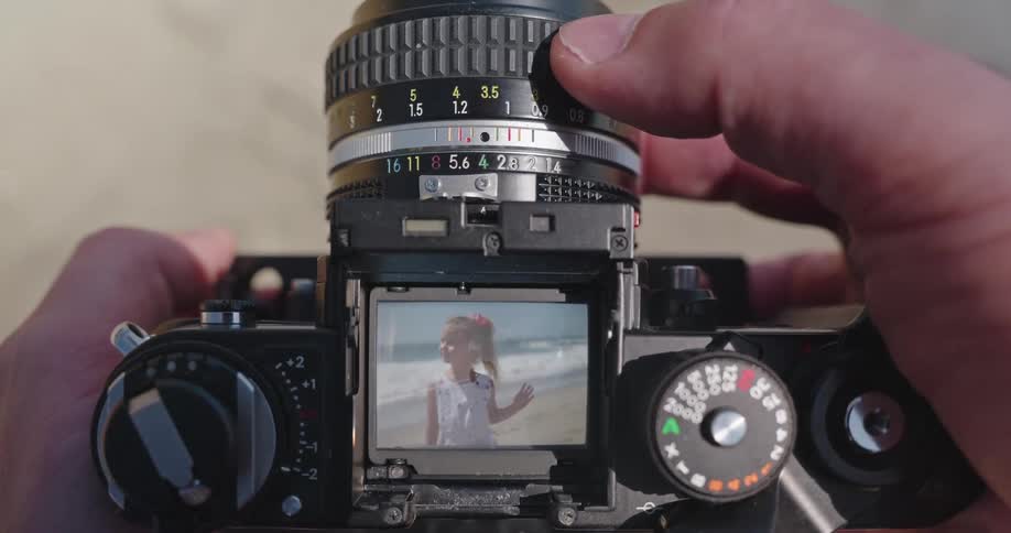 Close up point-of-view shot of a photographer focusing a vintage film camera on a little girl smiling on the beach
