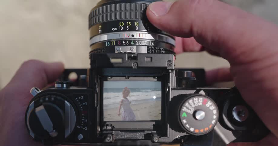 A photographer's point of view looking through a vintage waist-level viewfinder camera, focusing on a young girl playing on the beach by the ocean